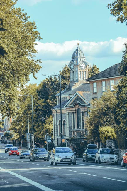 A large, historic stone building with ornate architectural details, including a central clock tower, decorative columns, and arched windows, situated on a street with a clear blue sky and scattered cl