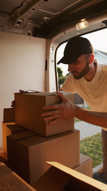Three young adults sit inside the open rear compartment of a light grey moving van during a home relocation, with visible cardboard boxes and a large plastic storage container. The woman on the left h