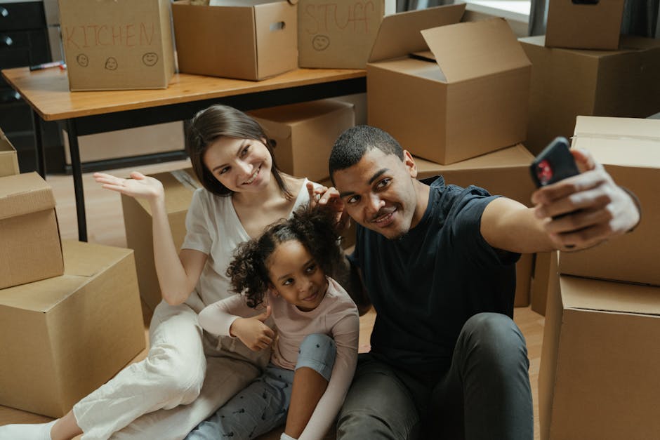 A family of three, including a woman with shoulder-length brown hair wearing a white t-shirt, a man with short dark hair wearing a black t-shirt, and a young girl with curly hair and wearing a pink long-sleeve top, taking a selfie together inside a home surrounded by numerous packed cardboard boxes and moving supplies. The boxes are of various sizes, some labeled with words like 'KITCHEN' and 'STUFF' and have smiley face drawings, and are stacked on furniture and the floor, with some placed on a wooden table in the background. The room is well-lit, with natural light coming from an unseen window, and the family appears happy and excited about the upcoming home relocation. The scene reflects the packing and moving process, with [COMPANY_NAME] providing house removals services to facilitate their furniture transport during their move to Grahame Park Estate in Colindale.