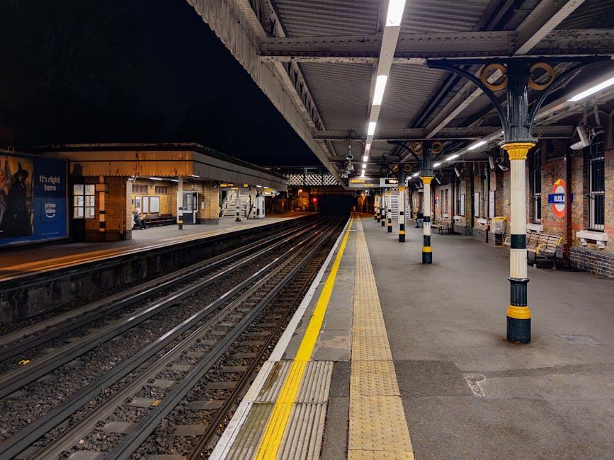 The image shows an empty train station platform at night, with multiple train tracks running parallel along the edge of the platform, which is made of concrete with tactile paving strips for accessibility. Support columns with black and yellow striped markings line the platform, supporting the overhead canopy that is illuminated by long fluorescent lights. A small seating area with benches and a shelter can be seen on the far side of the platform, while a staircase leading to an upper level or exit is visible in the background. A man seated on one of the benches is visible on the left side of the image, and a blue board with advertisements and station information is mounted on the brick wall behind him. The scene captures the typical urban environment of a busy railway station, where passengers wait and prepare for home relocation or other travel, with Man and Van Colindale occasionally involved in transportation and moving logistics.
