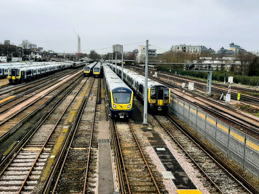 A view of multiple train carriages parked on parallel railway tracks at a railway station in an urban area, with some trains facing forward and others at slight angles. The trains are modern, with sleek yellow and grey fronts, and have large windows along their sides. The tracks are set on gravel beds, with several overhead power lines supported by metal poles stretching across the scene. Behind the railway yard, there are residential and commercial buildings, including multi-storey apartments and office blocks, with some trees and greenery visible in the background. The sky is overcast, casting diffuse light over the scene, capturing the busy atmosphere of a typical railway station used for passenger transport and home relocation logistics. Man and Van Colindale may use this train network for efficient furniture transport during house removals, supported by their professional packing and loading services.