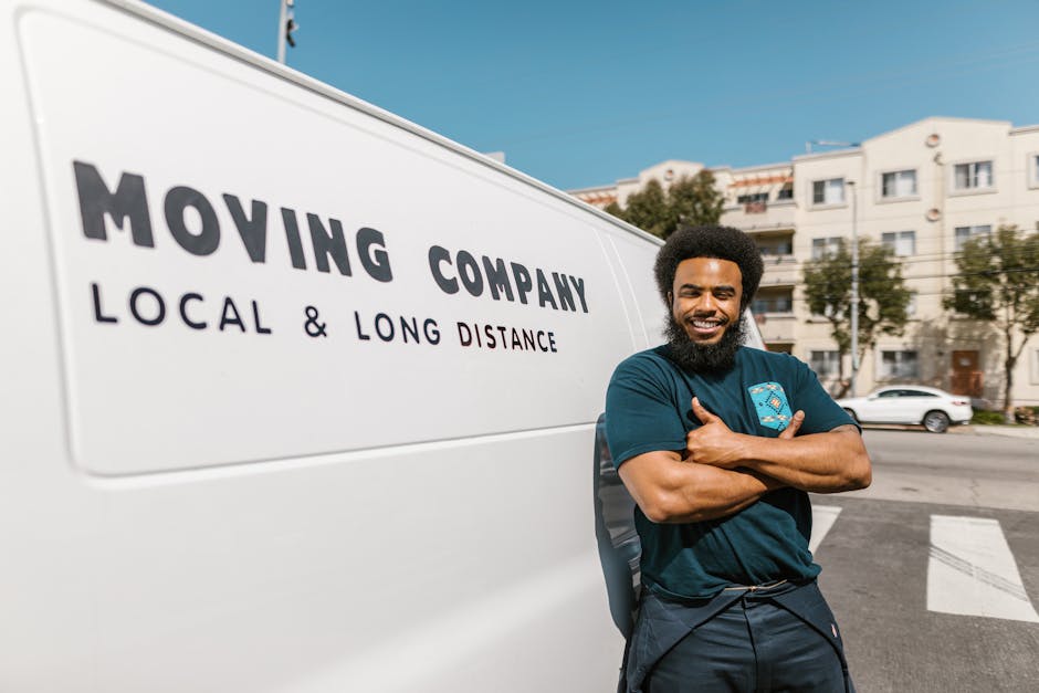 A man with a curly afro hairstyle and a full beard, casually dressed in a dark blue t-shirt, is standing next to a white moving van with 'MOVING COMPANY' and 'LOCAL & LONG DISTANCE' written on its sid