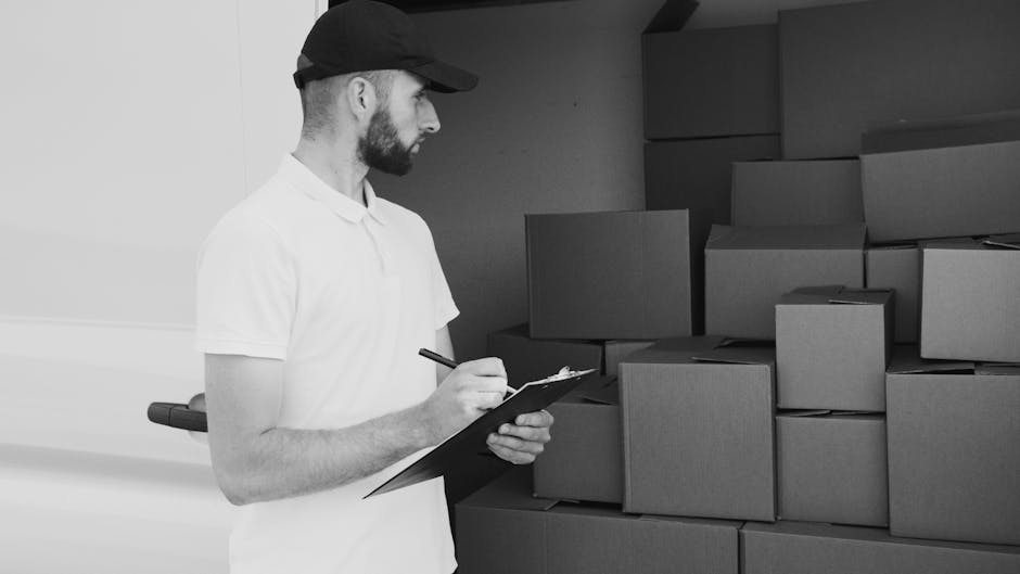 A man with a curly afro hairstyle and a full beard, casually dressed in a dark blue t-shirt, is standing next to a white moving van with 'MOVING COMPANY' and 'LOCAL & LONG DISTANCE' written on its sid