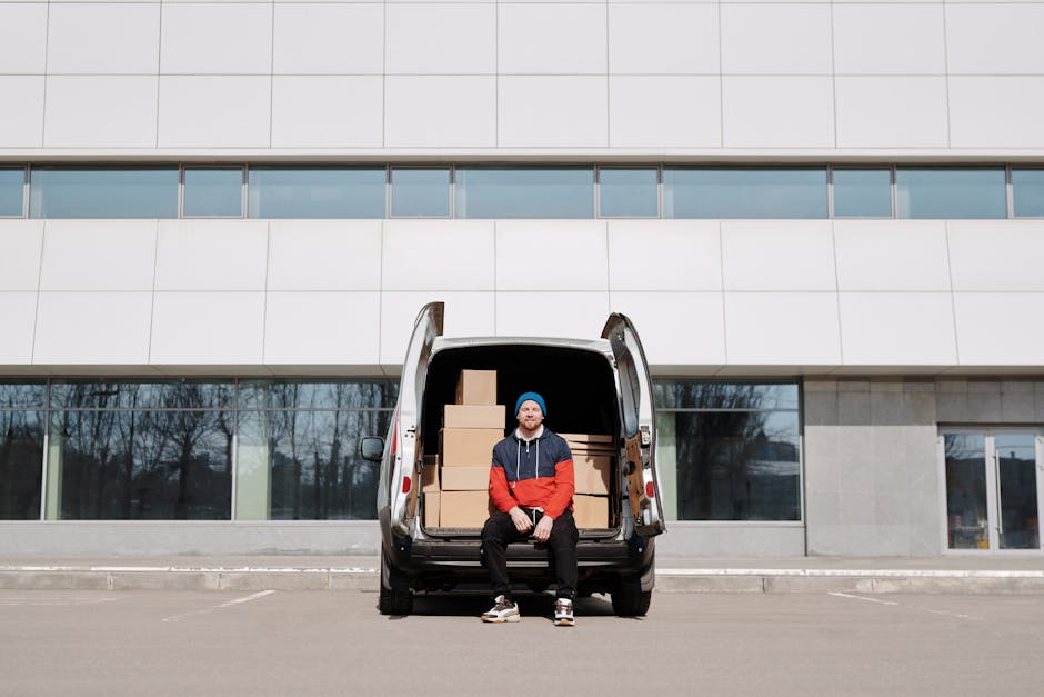 Two men sit on the open tailgate of a large black van used for furniture transport and house removals, parked outdoors on a paved surface. The man on the left wears a grey beanie, a green padded vest 