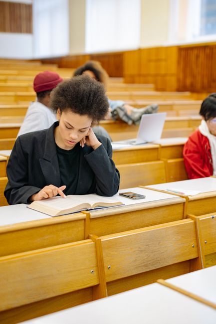 A group of students in a university lecture hall with wooden benches and a neutral wall background. One student with curly hair and a black hoodie is seated at a desk with an open laptop, holding a no